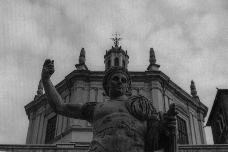 Low angle view of a historic marble statue with classical architecture in the background, set in an urban area. — Julius Caes